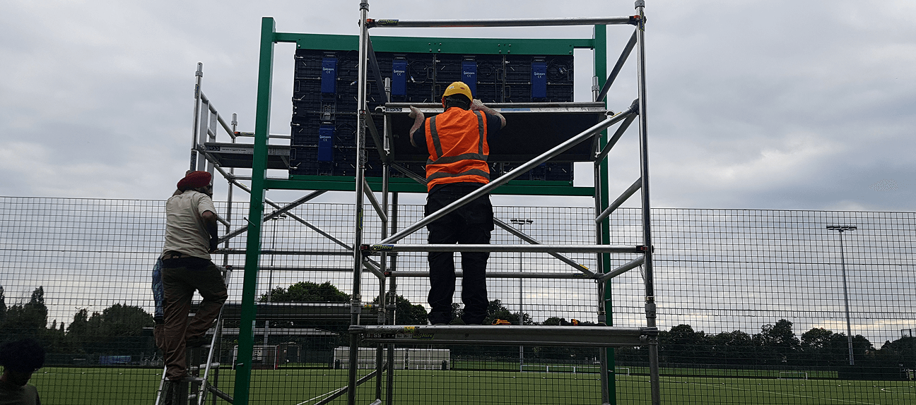 Technician installing LED scoreboard on steel frame structure.